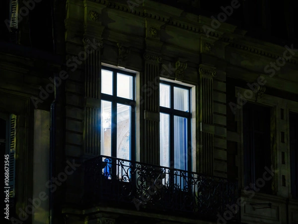 Fototapeta Illuminated window of a luxury apartment with ornate balcony railing at night, highlighting high-end real estate and elegant architectural design
