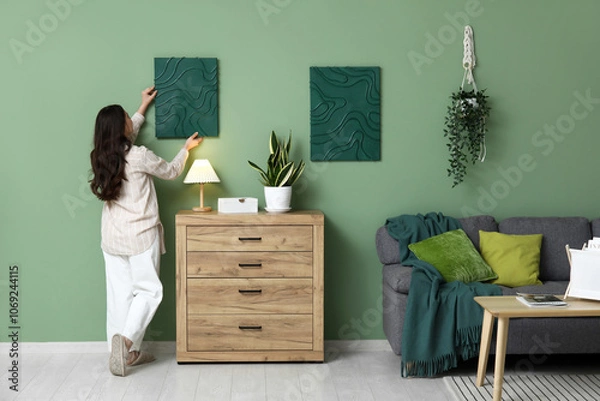 Fototapeta Young pretty woman hanging picture on green wall near chest of drawers in living room