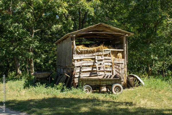 Fototapeta old wooden hayloft filled with hay against the background of green trees with wooden stairs and a wooden cart on a clear summer day