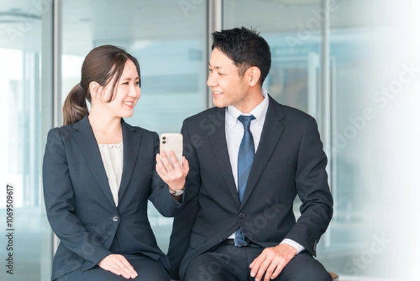 Fototapeta スマホ・スマートフォンを見て話すビジネスマンとビジネスウーマン（動画・アプリ・検索・ニュース）
