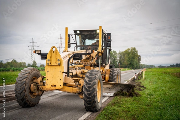 Fototapeta Motor grader spreading gravel at a construction site. Powerful road construction machinery for grading, leveling, and earthmoving. Ideal for infrastructure, civil engineering, and heavy projects. 