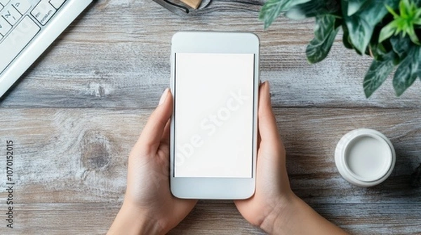 Fototapeta Hand holding a white cell phone with a blank screen on a desk in an office. Empty background space for advertisement, work, people, contact, marketing, and technology.