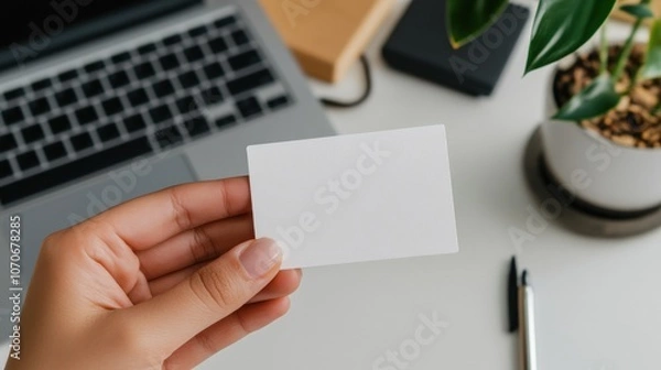 Obraz A hand displays a blank business card in a soft office environment featuring a laptop and a decorative plant. This setting suggests a professional context for networking or branding