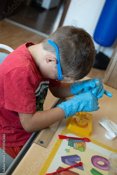Obraz Looking for a picture of a young boy doing a science experiment, wearing gloves and safety goggles, carefully using a dropper with colorful liquids. Something that shows kids learning in a fun, hands-