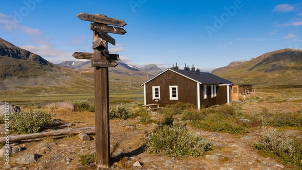 Fototapeta Wooden signpost and typical wooden nordic house provides shelter in remote wilderness for hikers in the scenic swedish mountains during sunny autumn day on Kungsleden trail. Lapland, Sweden