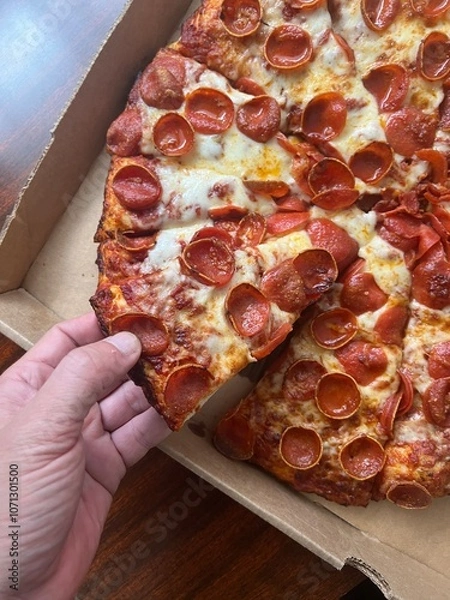 Fototapeta Hand taking a piece of cheesy large pepperoni pizza with cheese from a cardboard box. Takeout delivery to-go from a restaurant.