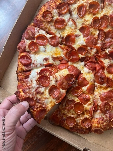 Obraz Hand taking a piece of cheesy large pepperoni pizza with cheese from a cardboard box. Takeout delivery to-go from a restaurant.