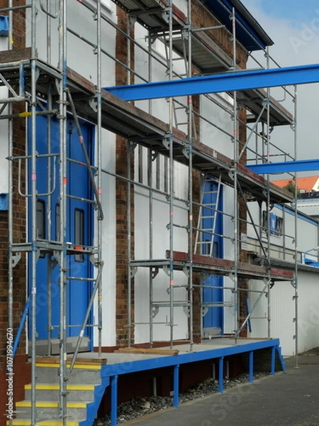 Fototapeta Scaffolding - with blue gate - The composition of the gray scaffolding in front of the blue industrial gate is an unusual perspective and gives the image a modern aesthetic.