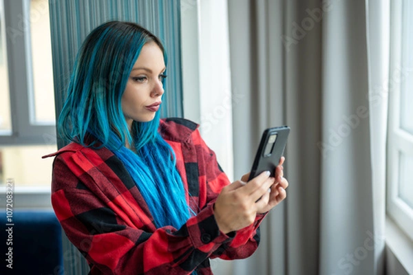 Fototapeta Portrait of beautiful blue haired caucasian hipster woman standing in front of window taking selfie with her mobile phone. Using technology.