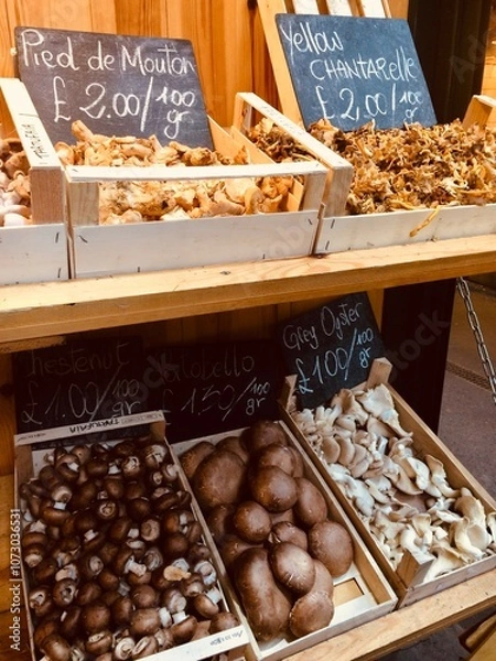 Obraz Various types of mushrooms in crates and displayed at a farmer’s market.  Chalkboard signs showing the names of the mushrooms and prices in British Pounds. 