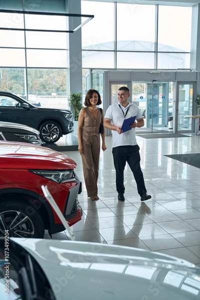 Fototapeta Salesman manager with file folder and female customer standing in car showroom