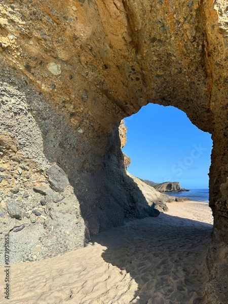 Obraz A captivating rock arch formation carved naturally over time, standing on a sandy beach under a bright blue sky.