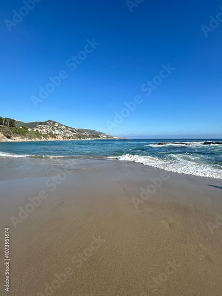Fototapeta A serene view of a pristine sandy beach stretching out towards the horizon, with gentle waves caressing the shoreline under a brilliant, cloudless blue sky.