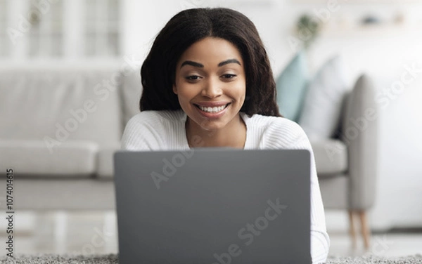 Fototapeta Remote job opportunities. Cheerful african american woman working distantly on laptop at home, lying on floor in living room interior. Smiling freelancer woman using pc computer