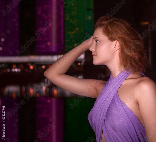 Fototapeta A young romantic girl against the background of modern buildings illuminated by multicolored LED light reflected in the water. Night city. Side view