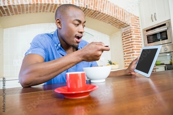 Fototapeta Young man having breakfast and using digital tablet