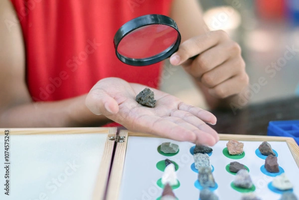 Fototapeta Asian schoolboy in red t-shirt is holding magnifying glass and looking natural rock on hand and comparing with the various sample rocks in box below, science study of rock concept.