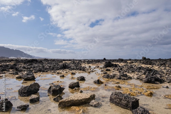 Fototapeta White sand beach with black rocks of volcanic origin. Mountains in the background and sky with big white clouds. Caletón Blanco, Lanzarote, Canary Islands, Spain