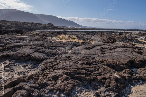 Fototapeta White sand beach with solidified volcanic lava flows, black rocks. Mountains in the background and sky with big white clouds. Caletón Blanco, Lanzarote, Canary Islands, Spain