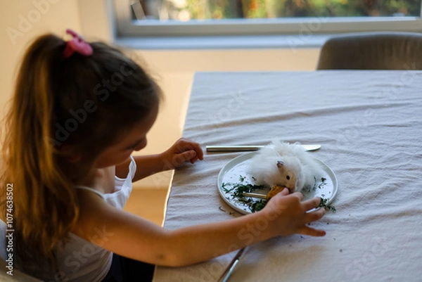 Fototapeta School ages girl feeds hamster at human plates and dining tables with fork and knife.