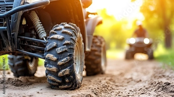 Fototapeta Close-up of an ATV tire on a dirt path, with a blurred rider in the background, illuminated by warm sunlight in a natural setting.