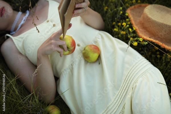 Obraz Woman,  lying in the green grass and reading a book, dressed in a beautiful yellow dress, holding an apple in the hand, enjoying rest.