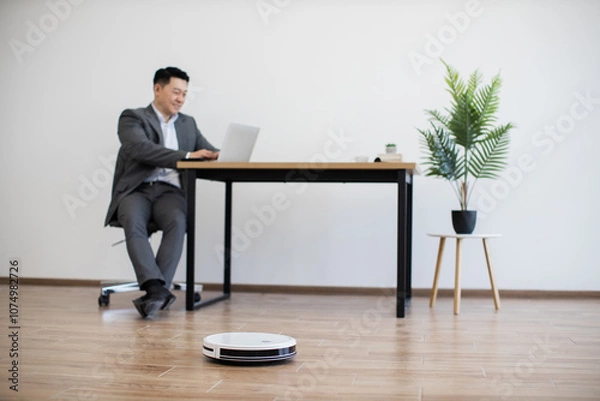 Fototapeta Asian businessman sitting at table using laptop in modern office space, while robot vacuum cleans floor. Scene depicts efficient, tech-savvy workspace with clean, minimalist design.