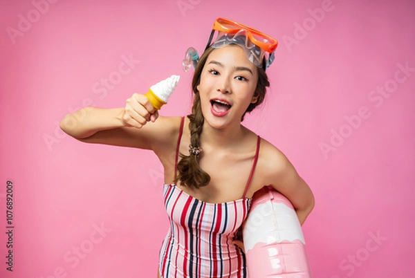 Obraz Cheerful woman in striped swimsuit holding inflatable swim ring and ice cream cone, wearing snorkel mask, standing against pink background