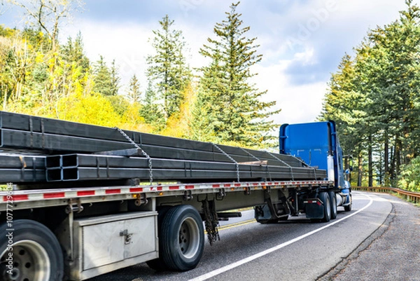 Obraz High cab blue big rig semi truck tractor transporting fastened by chains metal cargo on flat bed semi trailer running on the winding autumn highway road