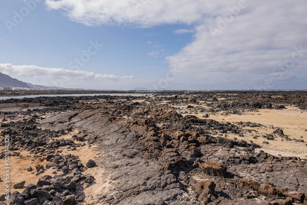 Fototapeta White sand beach with solidified volcanic lava flows, black rocks. Mountains in the background and sky with big white clouds. Caletón Blanco, Lanzarote, Canary Islands, Spain