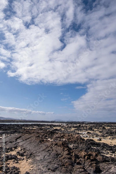 Fototapeta White sand beach with solidified volcanic lava flows, black rocks. Mountains in the background and sky with big white clouds. Caletón Blanco, Lanzarote, Canary Islands, Spain