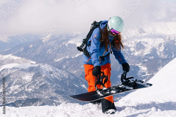 Fototapeta woman snowboarder wearing blue and orange ski suit and helmet  fastens snowboard bindings while standing on top of  slope in ski resort. Against background of grandiosely beautiful mountain range