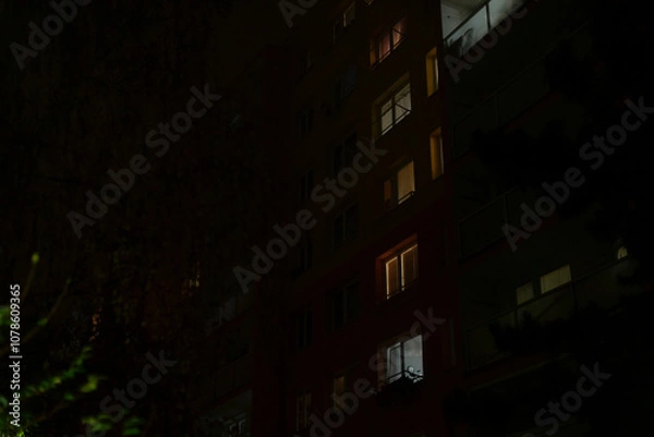 Fototapeta The scene presents a dark urban landscape, highlighting an apartment building adorned with illuminated windows that radiate light, creating a serene and quiet atmosphere during the tranquil nighttime