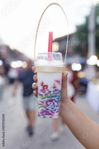 Obraz A hand holding a colorful Thai iced dessert in a clear plastic cup with a bamboo handle. The drink is layered with various colors and topped with coconut milk.