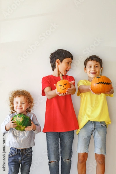 Obraz  Three real siblings standing together against white wall at cozy home and holding in their hands three orange and green pumpkins with scary faces, happy halloween at home