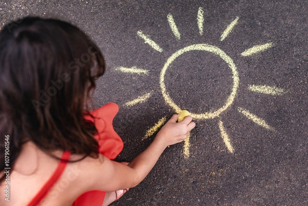 Obraz A little girl draws with colored chalk on the asphalt.
A drawing on a playground. A child draws a sun with chalk on an asphalt sidewalk.
