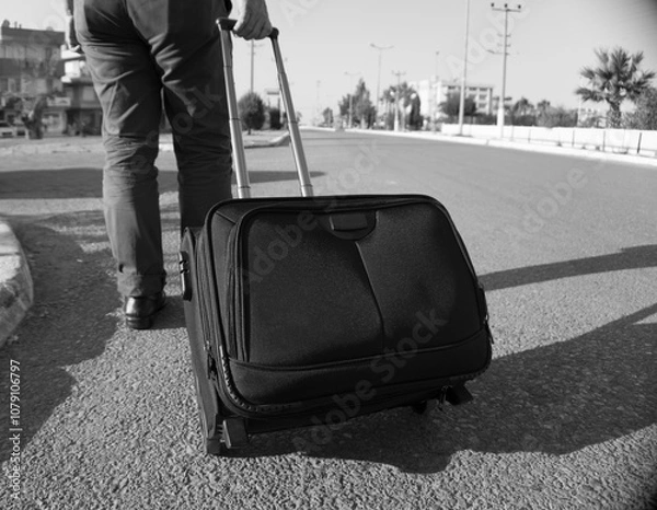Fototapeta A black and white image of a man pulling luggage on a road, representing travel or the journey of life.