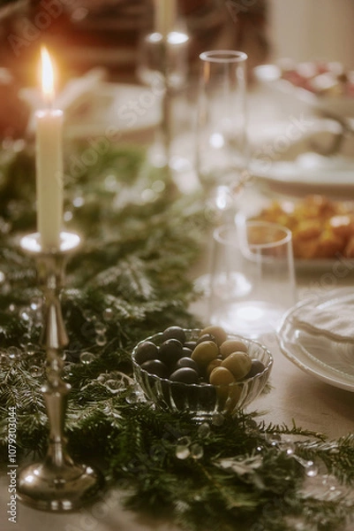 Fototapeta Vertical medium close up of olives in crystal bowl next to lit wax candle on table decorated with pine garland