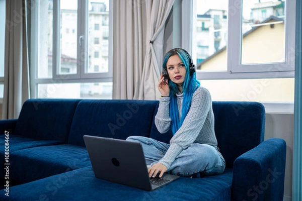 Fototapeta Beautiful blue haired hipster woman is working from home sitting on her couch, wearing headphones with microphone to attend a meeting with laptop. Concept of working from home, using technology.