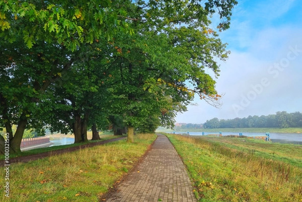 Fototapeta Serene Riverside Pathway in Lush Green Park Under Clear Blue Sky