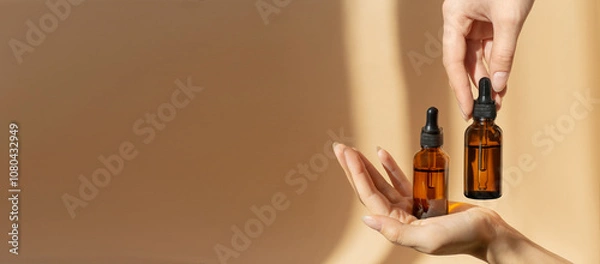 Obraz Women's hands hold bottles of amber glass with cosmetic serum on brown background. Mockup of container with dropper lid with collagen in rays of sunlight. Copy space,banner