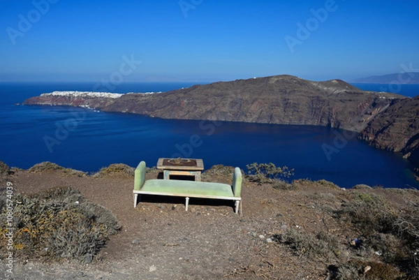 Obraz Eine Sitzbank mit Tisch steht am Wanderweg von Fira nach Oia am Wegesrand, und der Blick schweift über das blaue Mittelmeer auf die entfernte Stadt Oia.