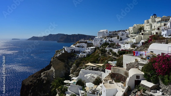 Obraz Ein wunderschönes Panorama der die Stadt Oia an der der Caldera von Santorin mit blick auf die Insel Thirasia. Man sieht auf die typischen weißen Häuser, blühende Pflanzen, bunte Wäsche auf der Leine.