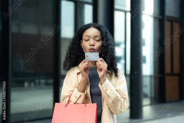 Obraz Stylish woman holding a credit card and shopping bag outdoors, expressing joy and satisfaction after a successful purchase.