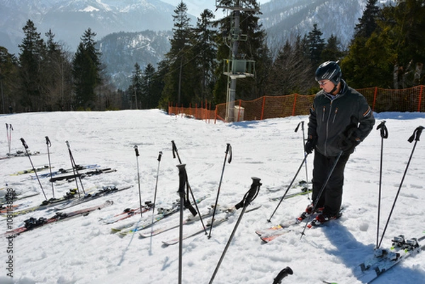 Fototapeta A skier is standing on a ski track next to laid out skis and poles of vacationers and is looking at these skis. Active winter recreation at a ski resort