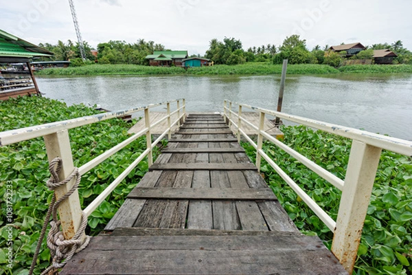 Fototapeta Rustic Wooden Pier Overlooking Riverside Scene with Water Hyacinths. It has white railings stretches over water and leading down to a peaceful riverside view.