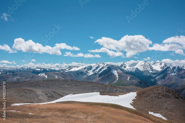 Fototapeta Aerial top view to stony pass and snow-white glacier on rocky hill in sunlight among high mountains in sunny day. Large colorful mountain range with snowy peaks in far away under clouds in blue sky.