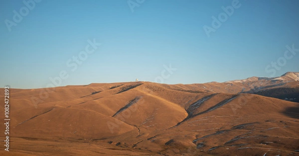 Fototapeta Golden hour light casting long shadows over undulating hills and dry grassland in a serene, high-altitude mountain landscape.