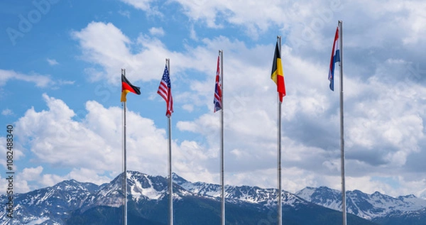 Fototapeta National flags of different countries flying in the wind against a blue sky and snowy mountain peaks.
