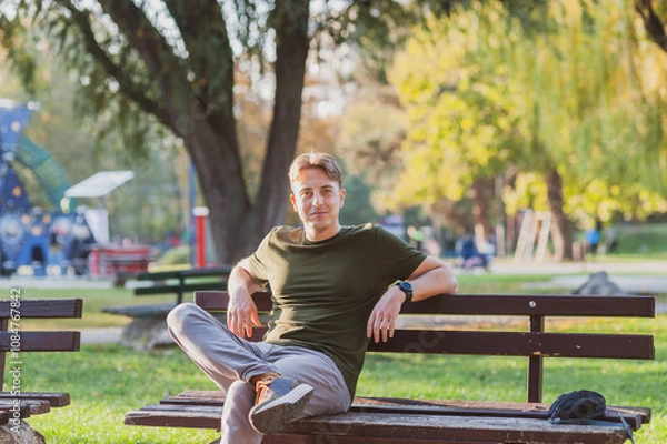 Obraz A cheerful young man is sitting alone in the park on the bench and enjoying the autumn sun